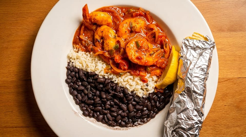 Shrimp creole with rice, black beans, lemon, and a foil-wrapped item on a white plate.