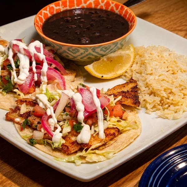 Two fish tacos with rice, black beans, and a lemon wedge on a white plate.