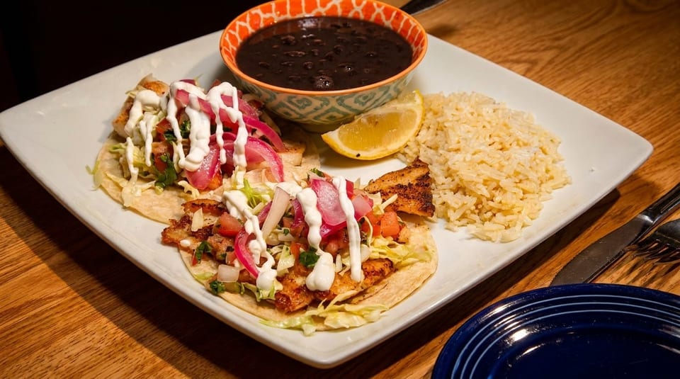 Two fish tacos with rice, black beans, and a lemon wedge on a white plate.