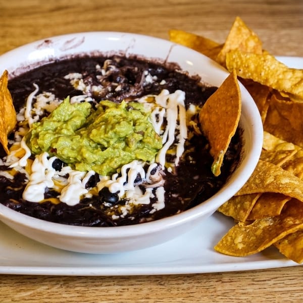 A bowl of black bean dip with guacamole, sour cream, and tortilla chips on a wooden table.