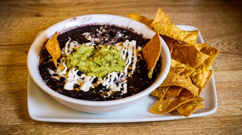 A bowl of black bean dip with guacamole, sour cream, and tortilla chips on a wooden table.