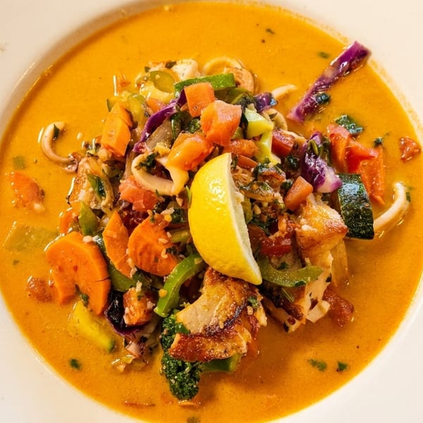A vibrant seafood stew with vegetables and a lemon wedge in a white bowl.