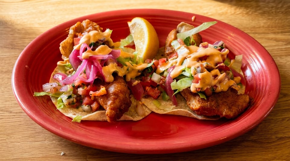 Two fish tacos with lettuce, pico de gallo, pickled onions, and sauce, served with a lemon wedge.
